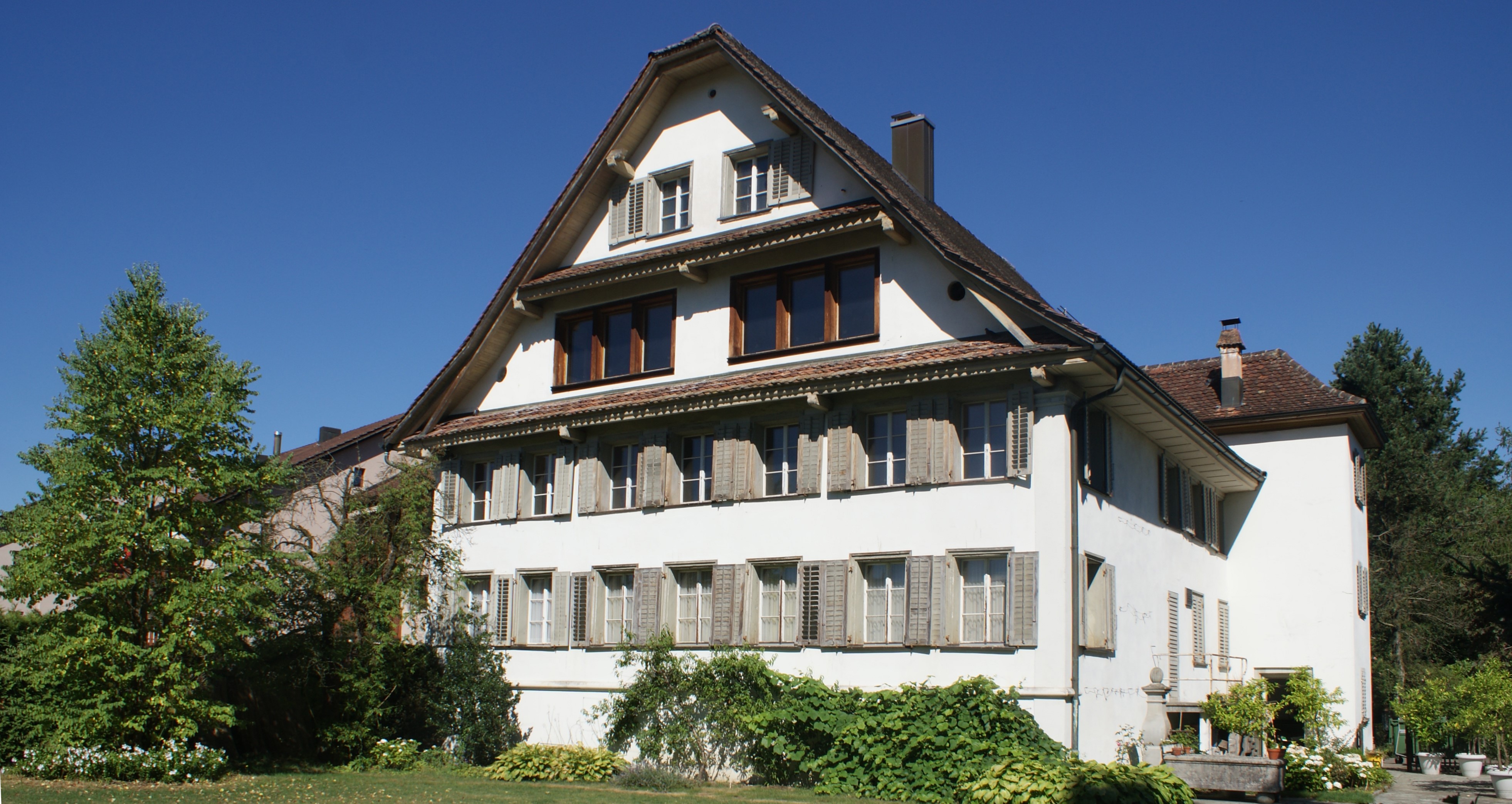 Ansicht der Fassade mit vielen Fenstern, im Vordergrund Garten, rechts Brunnen